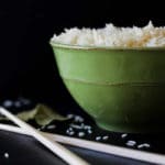Green bowl containing white rice, cross chopsticks and rice granules on black table.