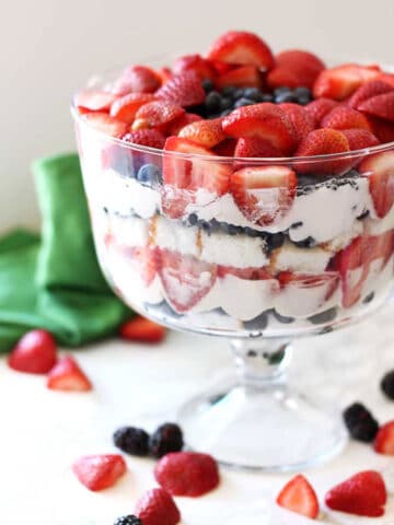 Trifle bowl containing strawberries, blueberries and cheesecake filling sitting on an white table, scattered berries.