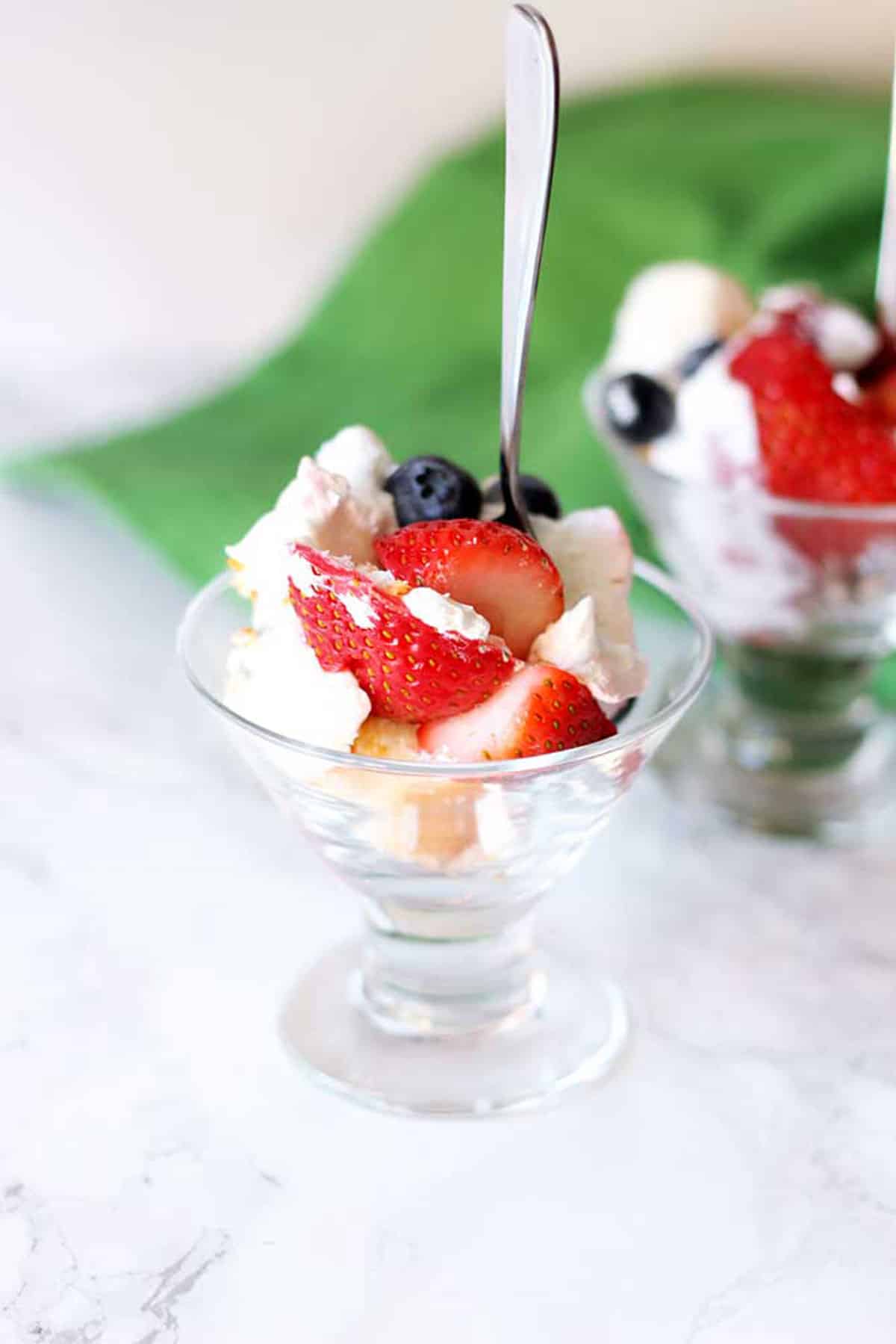 Parfait glass filled with strawberry, blueberry and angel foodmcake trifle with a spoon, green napkin in background.