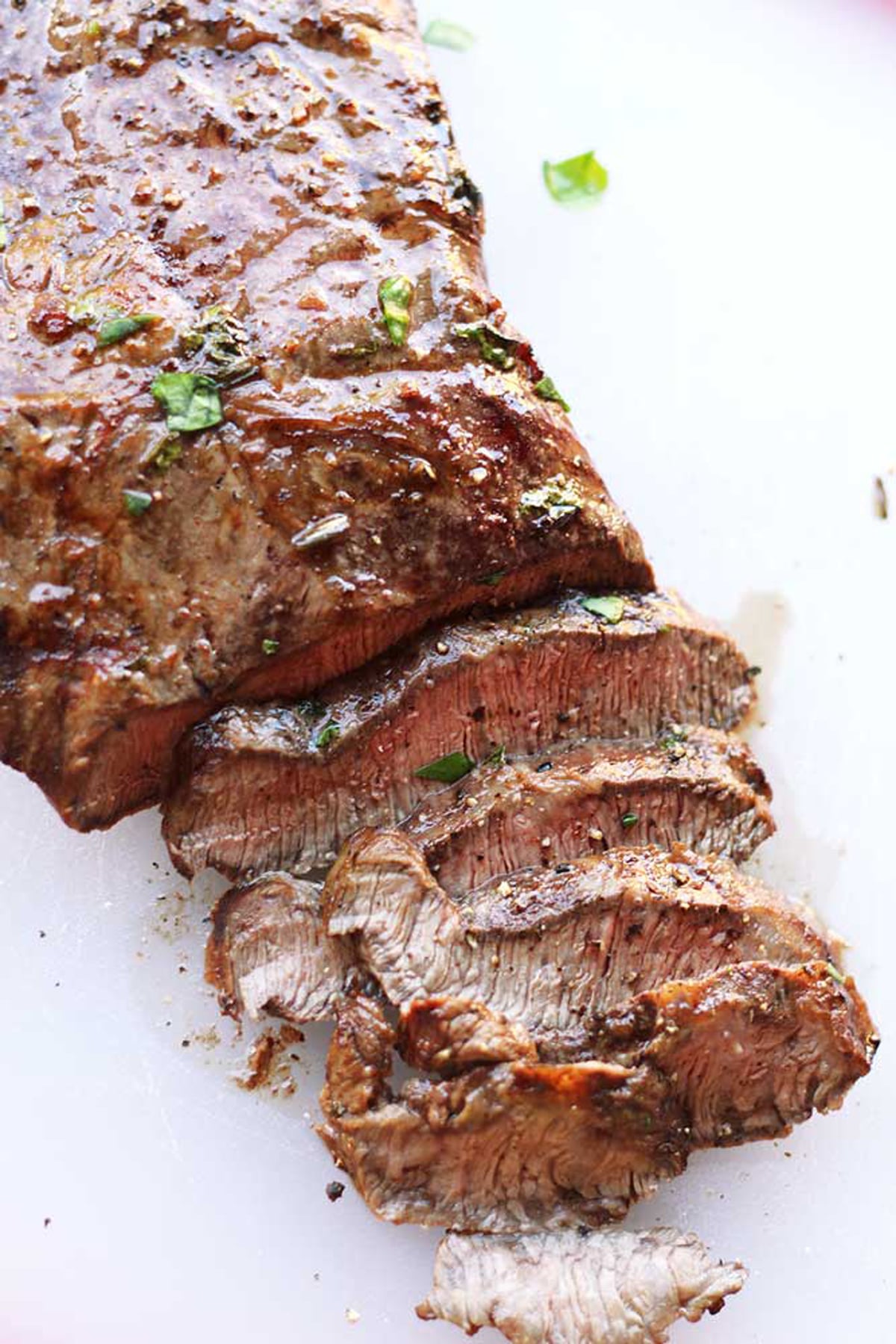 Sliced marinated flank steak sitting on a white table, topped with fresh parsley and fresh ground pepper.