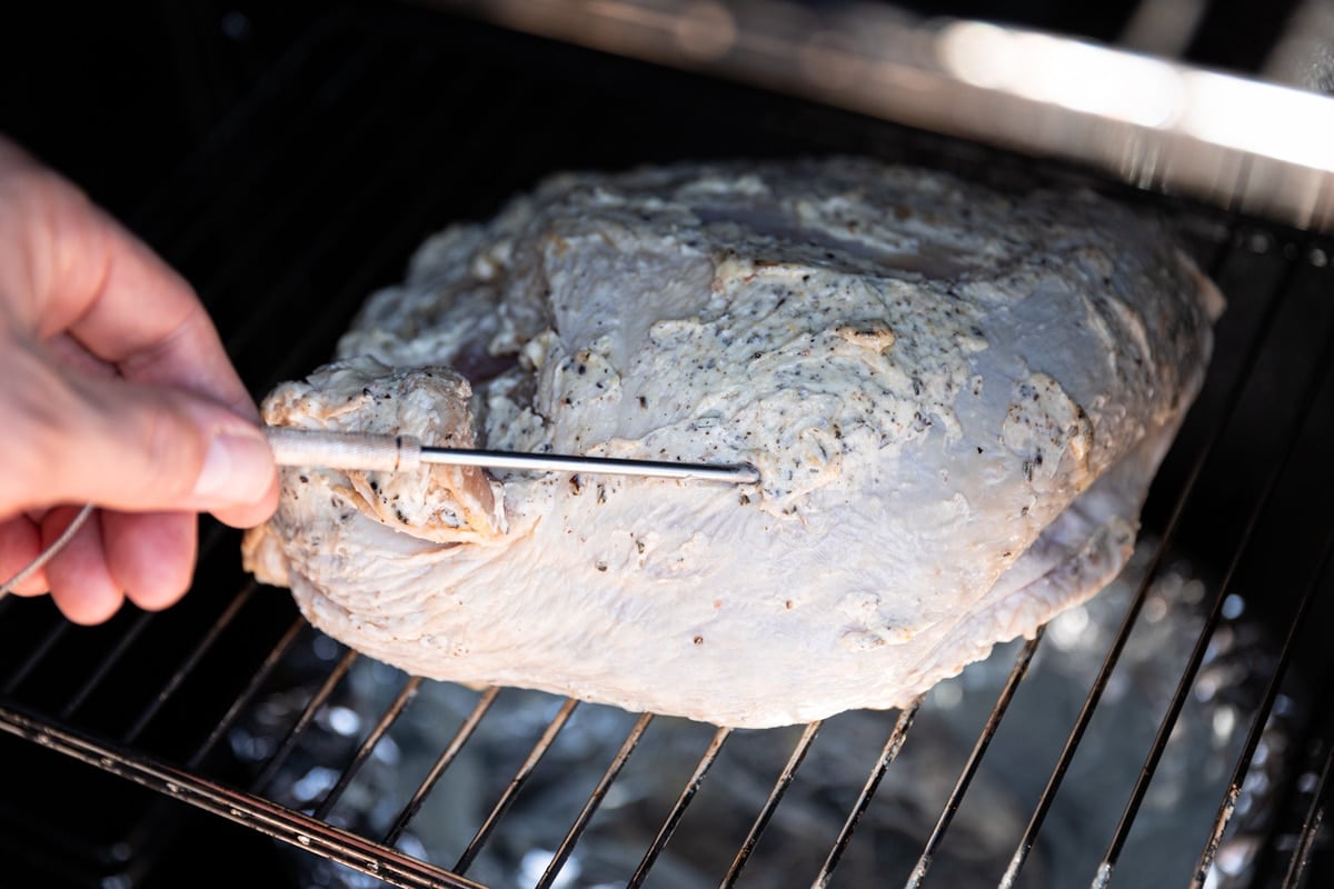 Person inserting a temperature probe into a raw turkey breast on a smoker. 