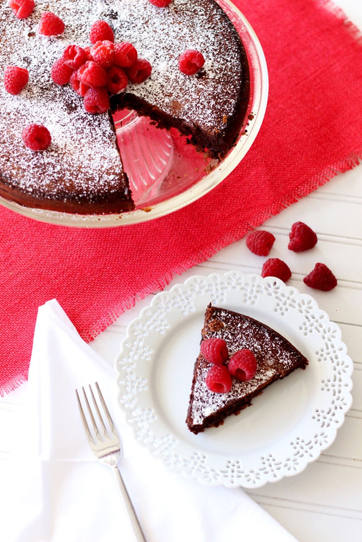 A slice of Flourless Chocolate Torte topped with powdered sugar and raspberries sitting on a plate, fork and remaining cake sitting on a white table with red napkin.
