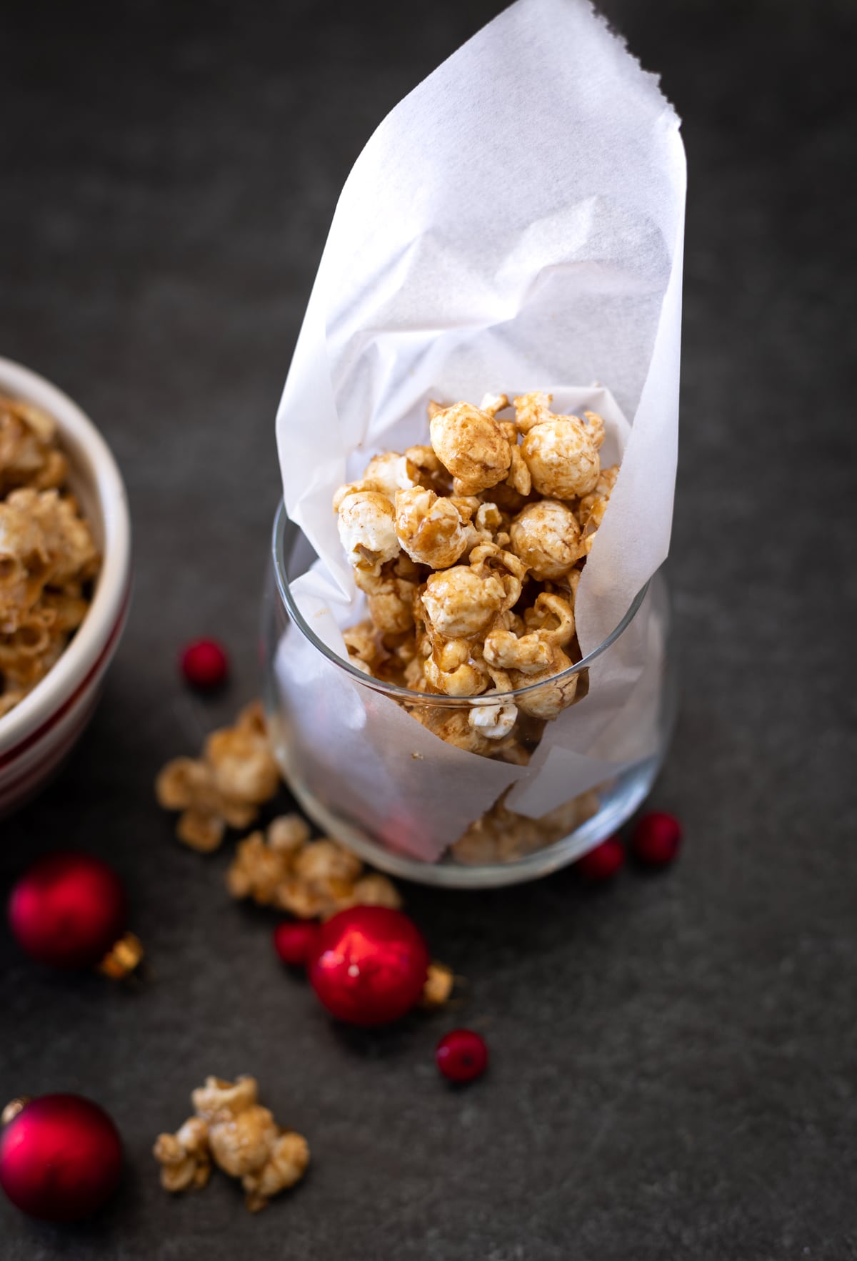 Short glass with white tissue containing Bourbon Caramel Popcorn, red Christmas balls and bowl of popcorn on black table. White tissue paper in glass.