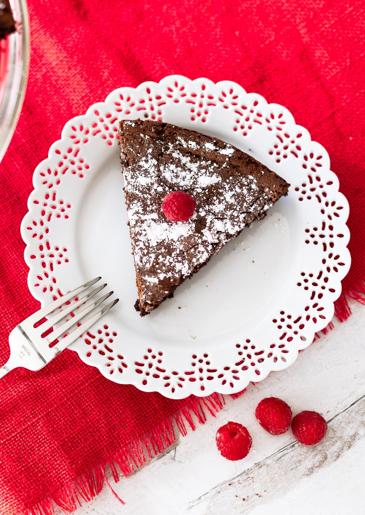 A slice of Flourless Chocolate Torte topped with powdered sugar and raspberries on a white lace plate, silver fork on side.