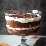 Trifle bowl filled with chocolate pudding, ladyfingers, and whip cream on a table.