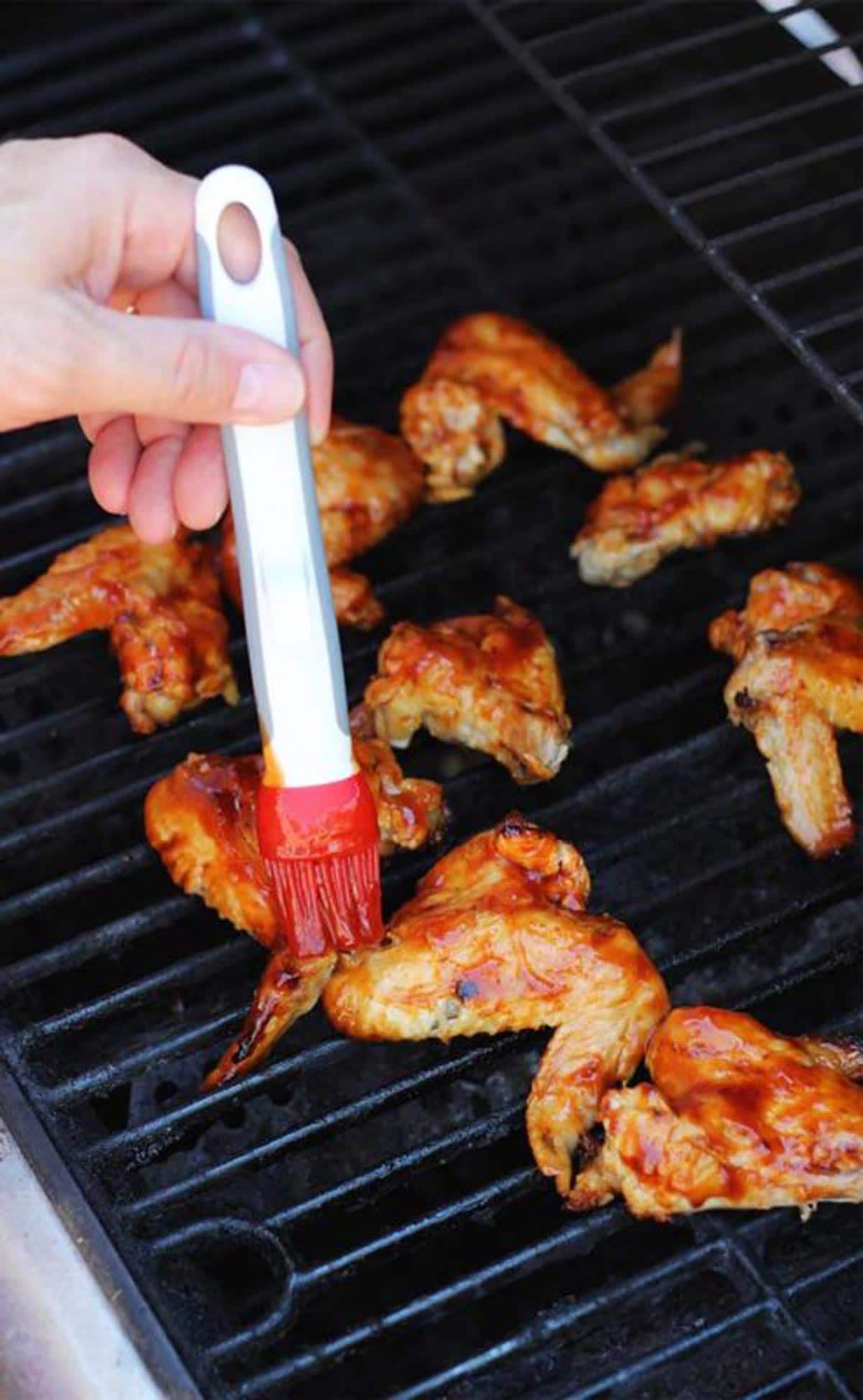 9 BBQ Chicken Wings on a grill, hand brushing BBQ sauce on a chicken wing while grilling.