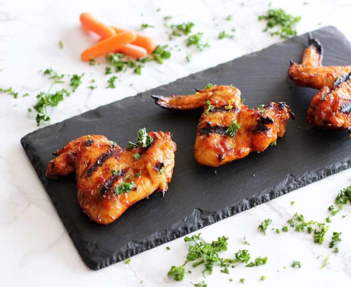 Black slate plate featuring 3 BBQ chicken wings topped with parsley, sitting on a white table containing scattered carrots and fresh parsley.