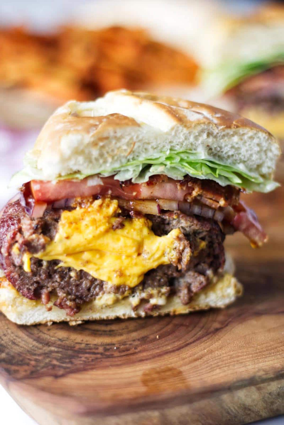 A burger cross section, sitting on a wood cutting board, cheese spilling out the middle with bacon, onion, tomato, lettuce.