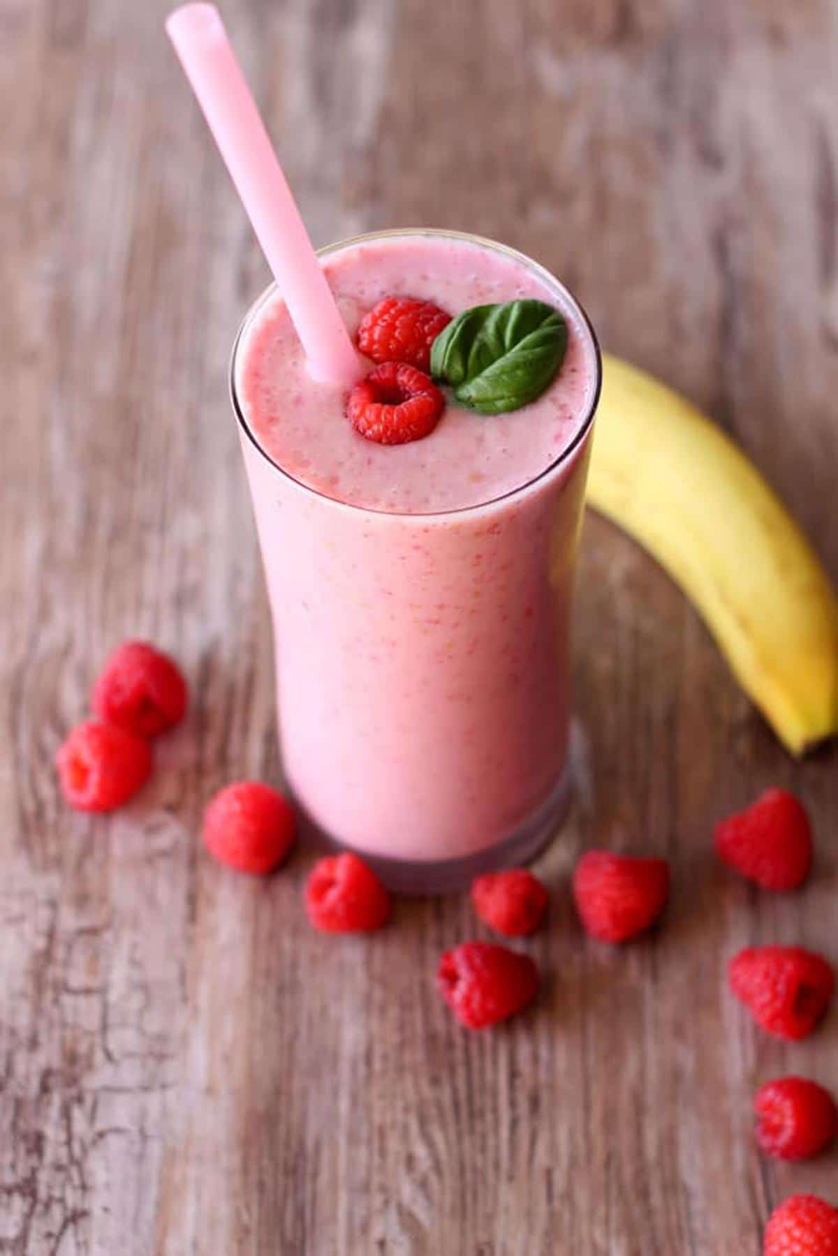 Glass containing a berry smoothie topped with 2 raspberries and a mint spring, berries and banana on a wooden table.