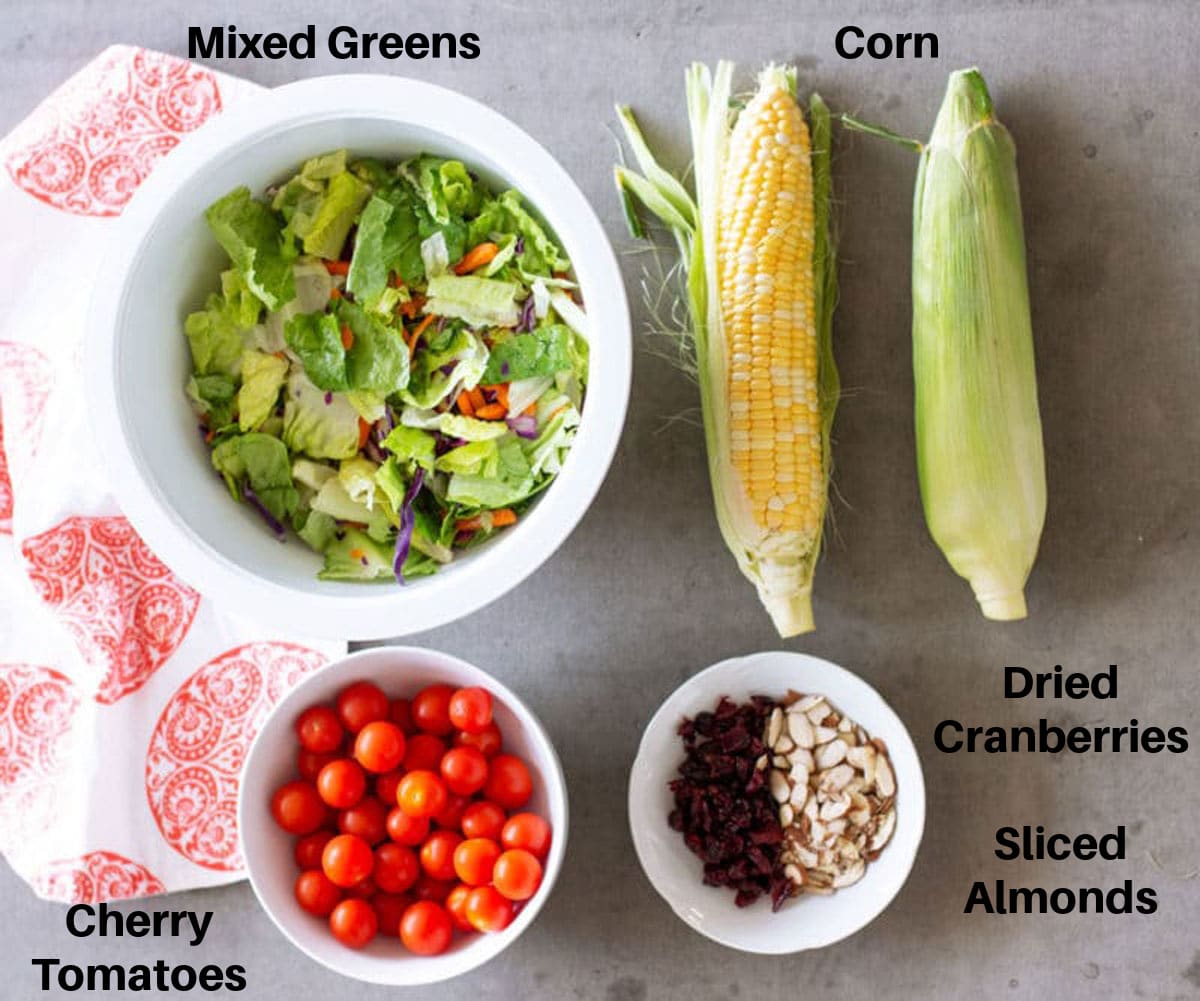 Bowl of lettuce, 2 ears of corn, cherry tomatoes, almonds, and cranberries on counter.