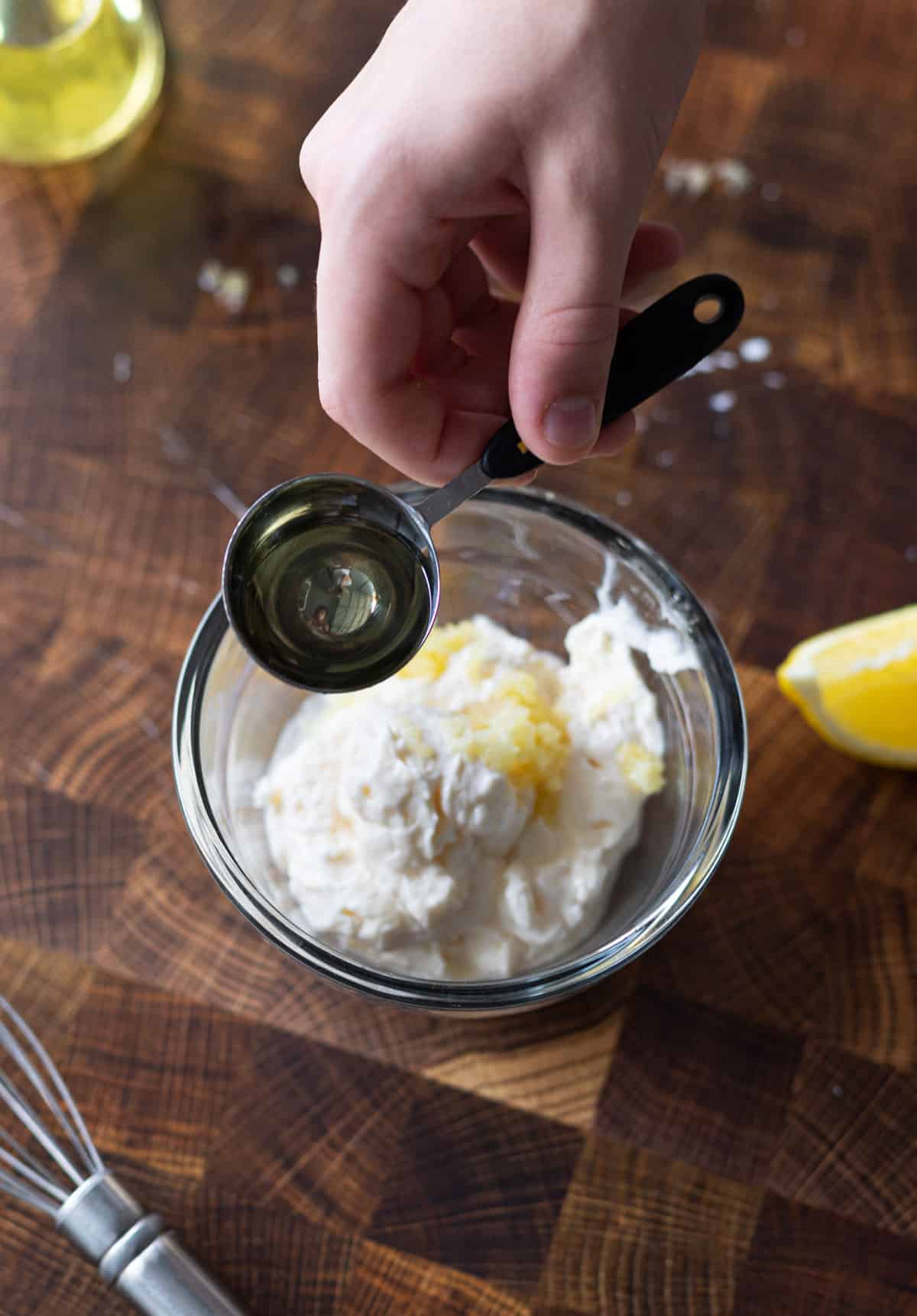 A tablespoon of olive oil being poured into a small bowl of mayonnaise and garlic.