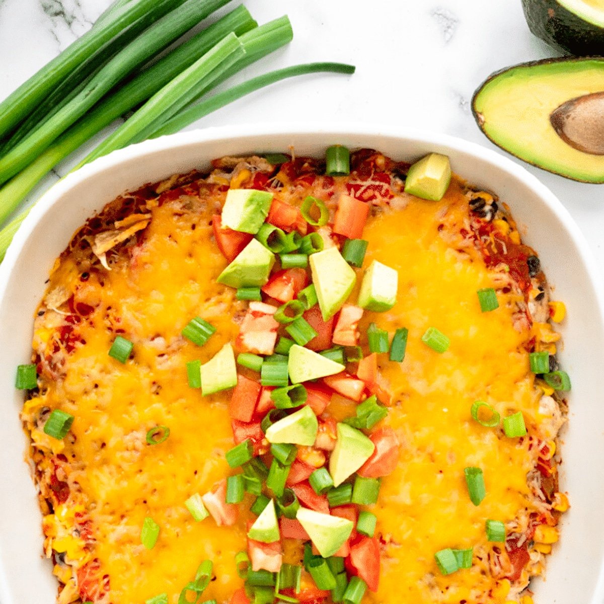 White casserole dish containing mexican casserole topped with green onions, tomato, and avocado.