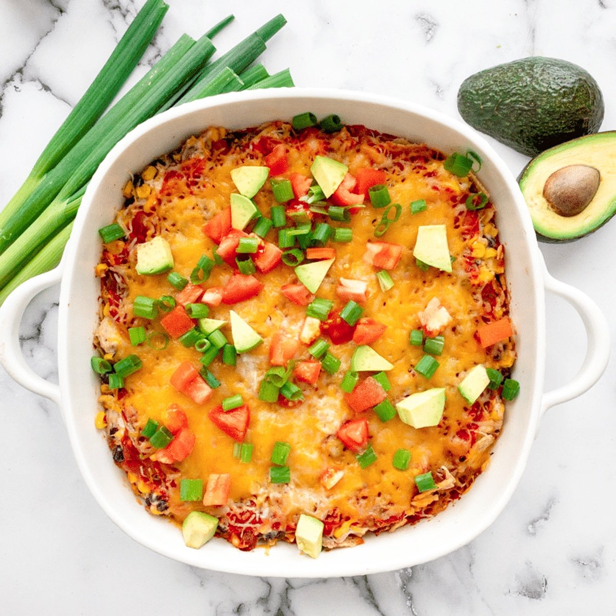 Mexican Chicken casserole in white dish on counter topped with tomatoes and avocado. 