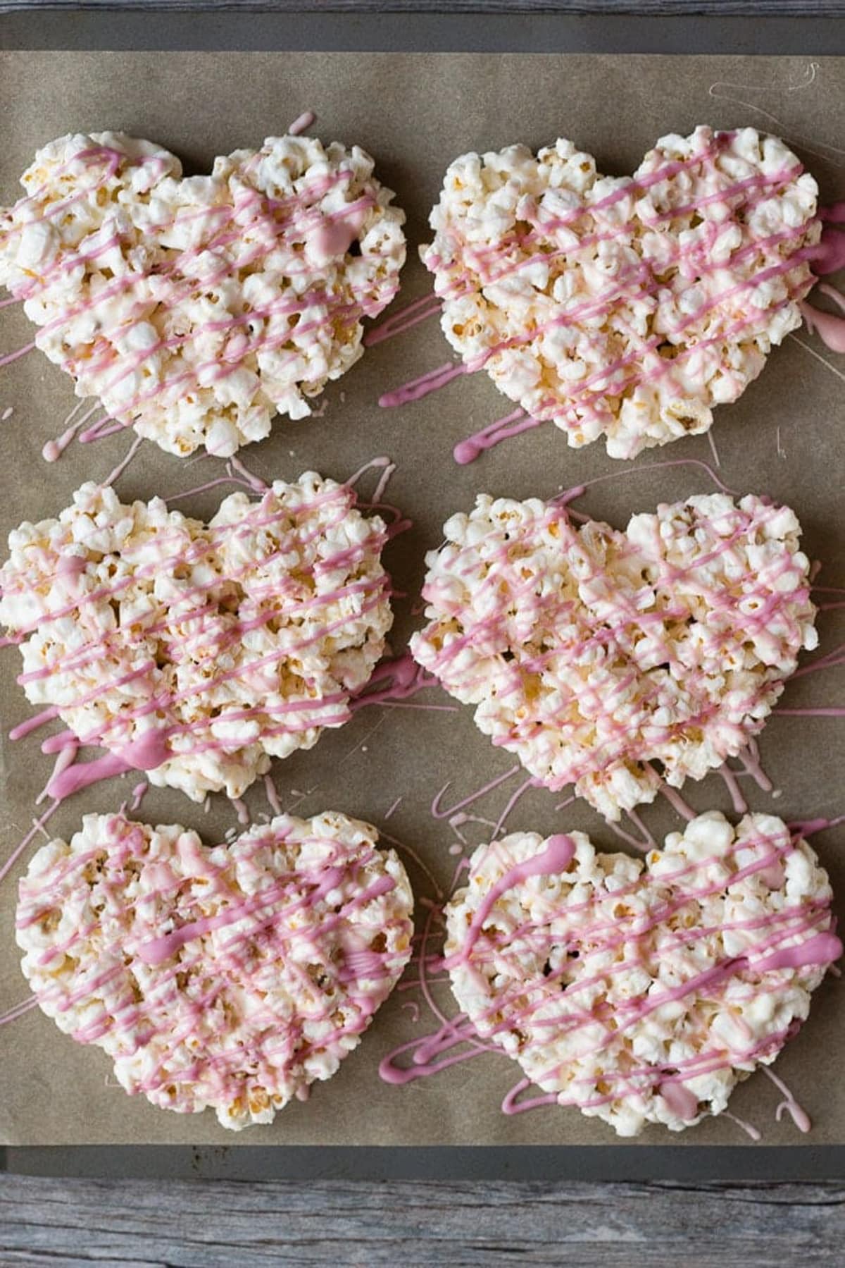 Six marshmallow popcorn hearts on a parchment-lined cookie sheet.