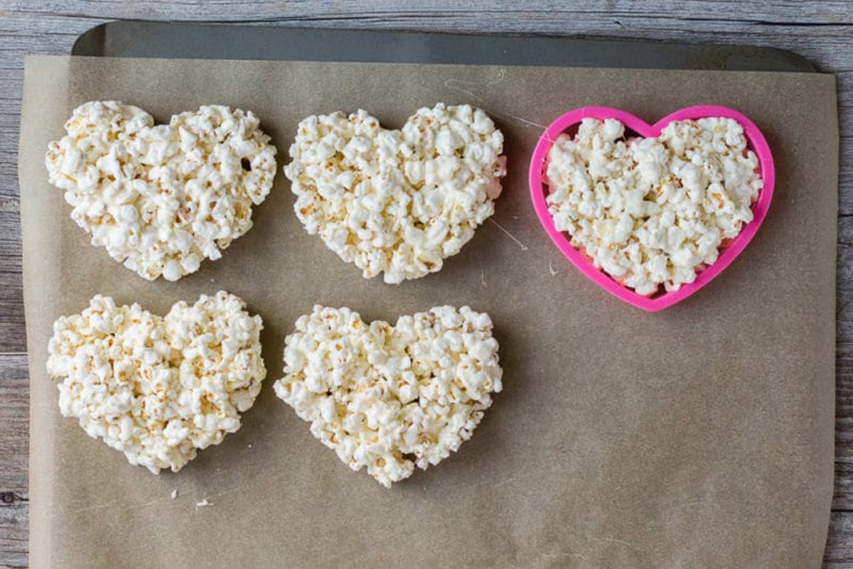 Marshmallow popcorn hearts being made on a parchment paper lined cookie sheet with a pink cookie cutter.