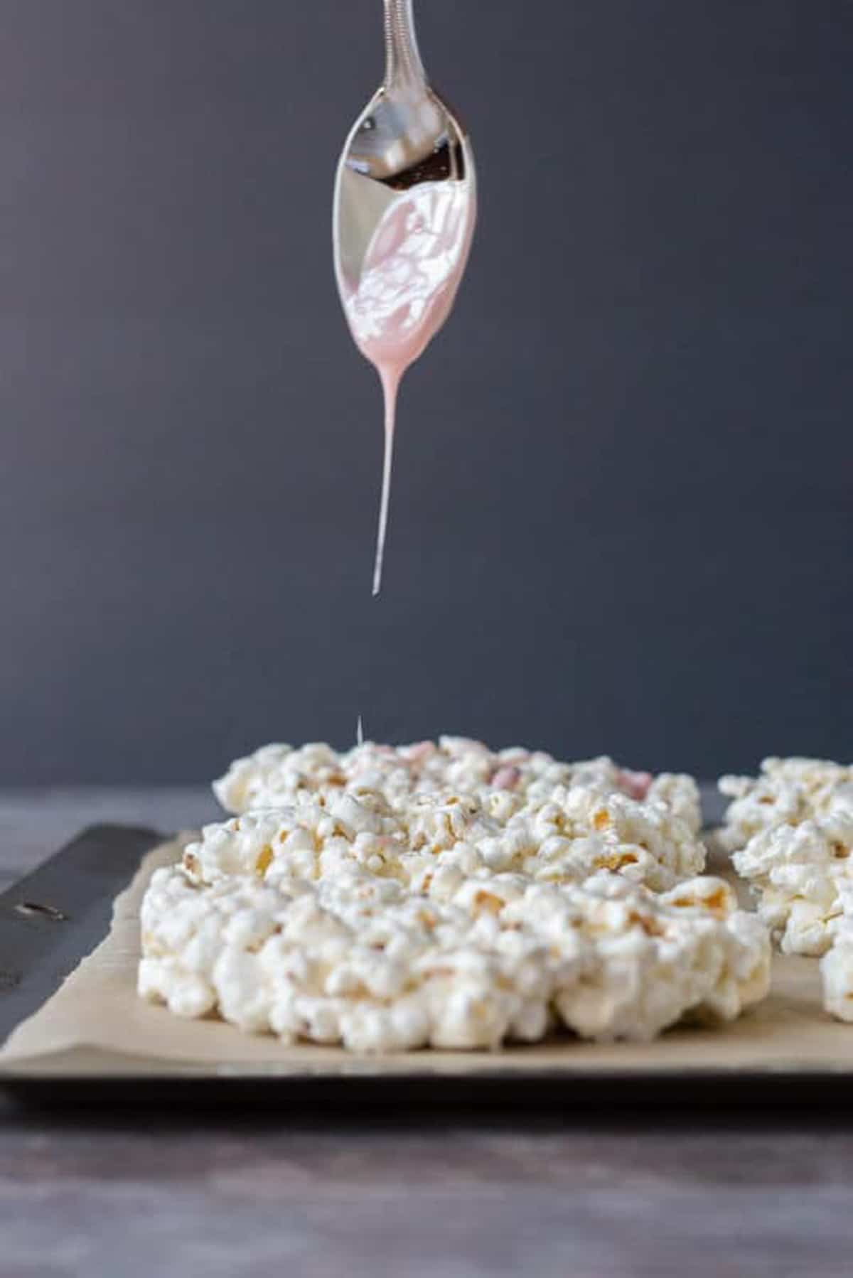 Pink colored white chocolate dripping off a silver spoon onto marshmallow popcorn hearts.