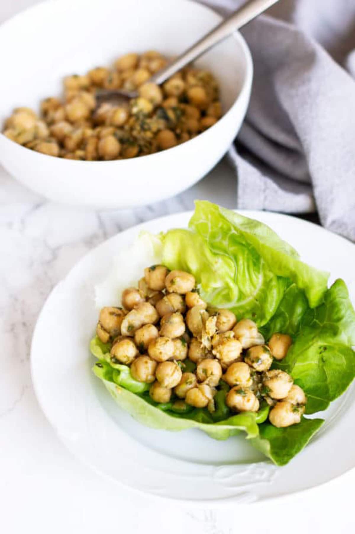 White plate containing a Vegetarian lettuce wrap, bowl of pesto chickpeas on table.