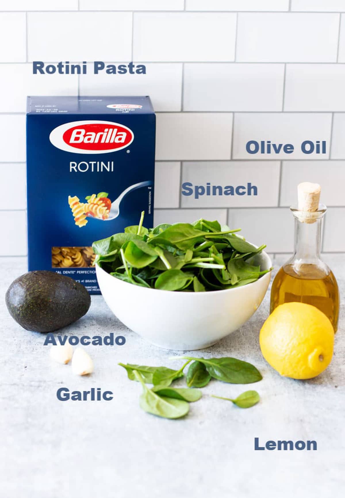 Rotini pasta, spinach, olive oil, lemon, garlic and avocado on a counter.