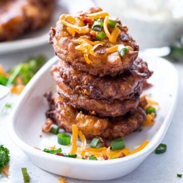 Stacked fried corn fritters in a dish topped with bacon, scallions, and cheese.