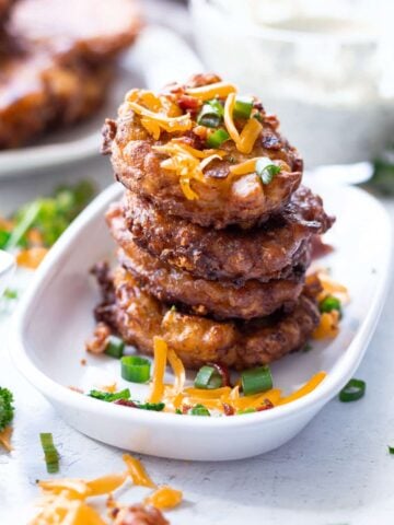 Stacked fried corn fritters in a dish topped with bacon, scallions, and cheese.