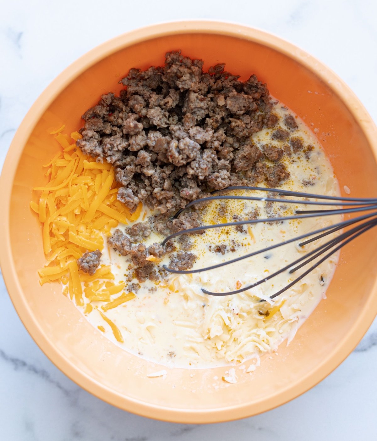 Person adding in grated cheese and cooked sausage to bowl of liquid casserole ingredients. 
